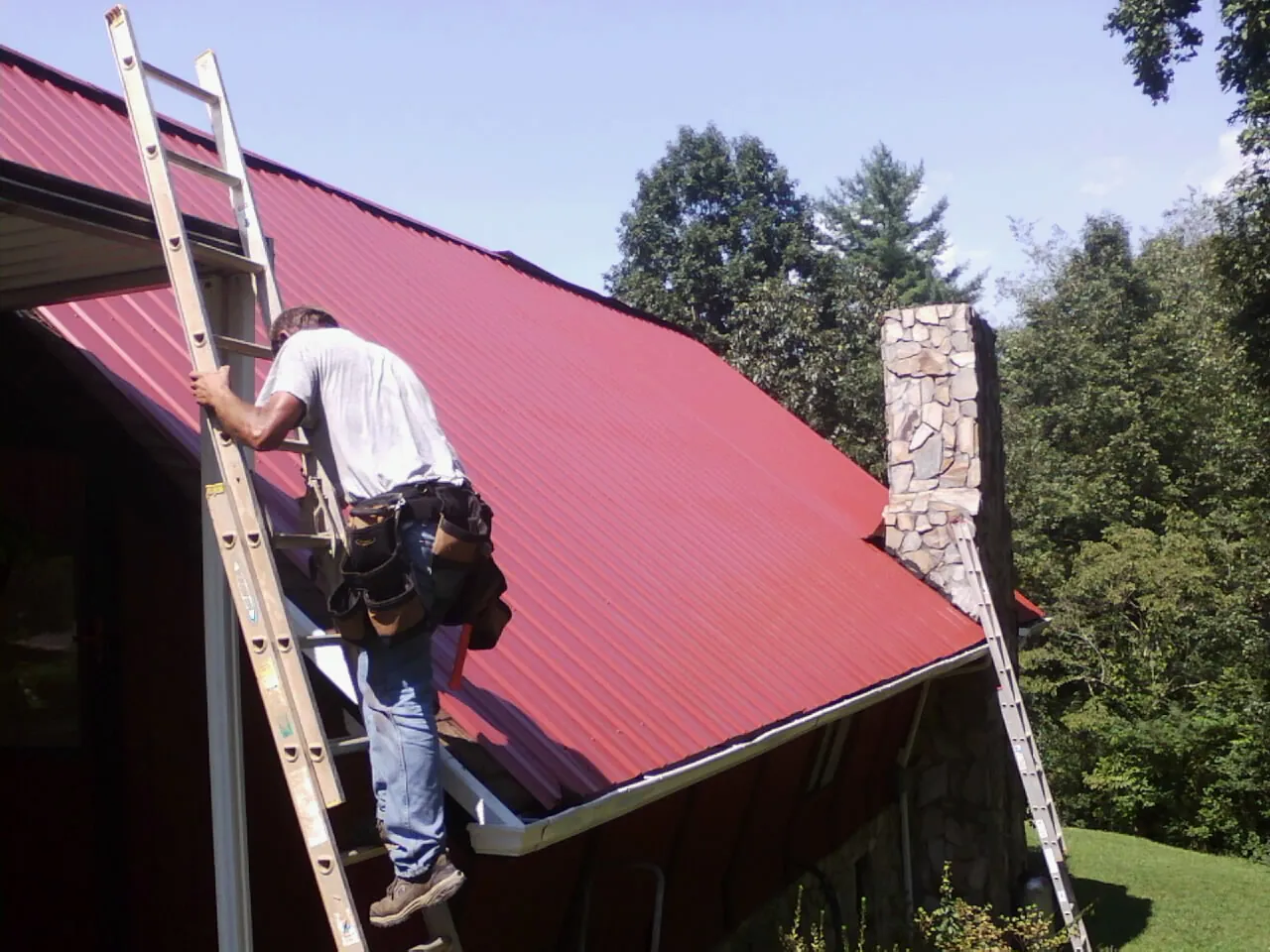 Roofer on a ladder beside a red metal roof and stone chimney — Soffit & Fascia Repair by Roofing Brothers in Eufaula